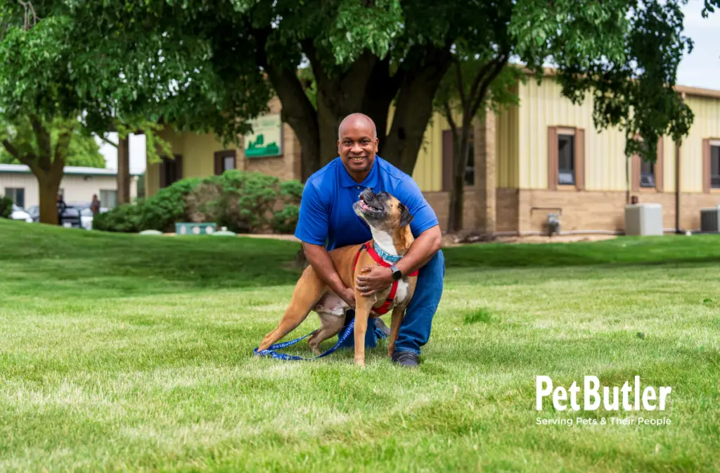 Man kneeling on a grassy lawn smiling while holding a happy dog on a leash.