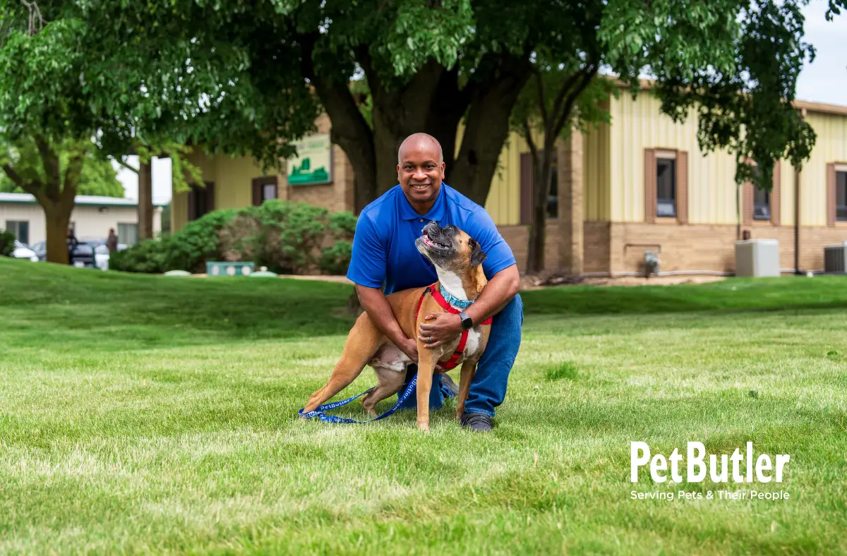 Man kneeling on a grassy lawn smiling while holding a happy dog on a leash.